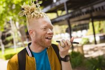 Glücklicher Albino-Afrikaner mit Dreadlocks im Park, der mit dem Smartphone spricht. digitaler Nomade unterwegs, unterwegs in der Stadt. — Stockfoto