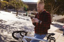 Усміхнений альбіноамериканський чоловік з дредлоками на велосипеді за допомогою смартфона. цифровий кочівник на ходу, вихід і близько в місті . — стокове фото