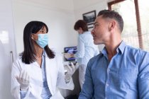 Dentista biracial usando máscara facial e conversando com paciente do sexo masculino na clínica odontológica moderna. serviços de saúde e odontologia. — Fotografia de Stock