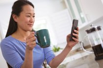 Mujer asiática feliz bebiendo café y usando teléfono inteligente en la cocina. estilo de vida y relajarse en casa con la tecnología. - foto de stock