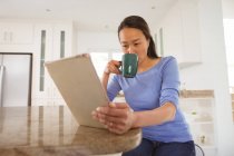 Konzentrierte asiatische Frau, die Kaffee trinkt und Tabletten in der Küche benutzt. Lifestyle und Entspannung zu Hause mit Technologie. — Stockfoto