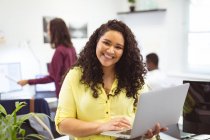 Portrait of smiling biracial businesswoman using laptop looking at camera in modern office. business and office workplace. — Stock Photo