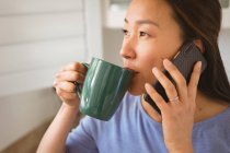 Retrato de mujer asiática feliz bebiendo café y usando smartphone en la cocina. estilo de vida y relajarse en casa con la tecnología. - foto de stock