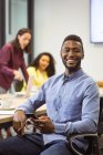 Porträt eines lächelnden afrikanisch-amerikanischen Geschäftsmannes, der im modernen Büro mit dem Smartphone in die Kamera blickt. Geschäfts- und Büroarbeitsplätze. — Stockfoto