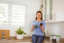 Glückliche asiatische Frau, die Kaffee trinkt und ihr Smartphone in der Küche benutzt. Lifestyle und Entspannung zu Hause mit Technologie. — Stockfoto