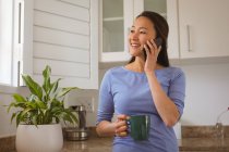 Счастливая азиатка смотрит в окно, пьет кофе и пользуется смартфоном на кухне. стиль жизни и отдых дома с технологией. — стоковое фото