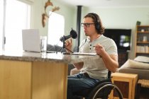 Kaukasischer behinderter Mann, der einen Podcast mit einem Mikrofon aufzeichnet, der zu Hause im Rollstuhl sitzt. Blogging, Podcast und Sendetechnologie-Konzept — Stockfoto