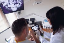 Odontóloga Biracial usando máscara facial examinando los dientes de un paciente masculino en una clínica dental moderna. negocio de salud y odontología. - foto de stock