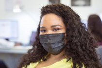 Retrato de mulher de negócios birracial sorridente usando máscara facial olhando para a câmera no escritório moderno. negócio e escritório local de trabalho durante a pandemia covid 19. — Fotografia de Stock