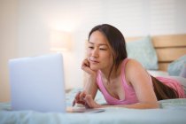 Pensativo mujer asiática acostada en la cama, utilizando el ordenador portátil. estilo de vida y relajarse en casa con la tecnología. - foto de stock