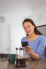 Felice donna asiatica bere caffè e utilizzando smartphone in cucina. stile di vita e relax a casa con la tecnologia. — Foto stock