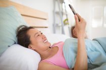 Mujer asiática feliz acostada en la cama, descansando y usando un teléfono inteligente. relajarse en casa con la tecnología. - foto de stock