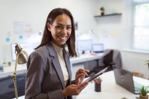 Portrait d'une femme d'affaires biraciale souriante utilisant une tablette regardant la caméra dans un bureau moderne. lieu de travail professionnel et de bureau. — Photo de stock