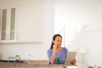 Mujer asiática sentada en la mesa, tomando café y usando la tableta en la cocina. estilo de vida y relajarse en casa con la tecnología. - foto de stock