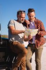 Happy caucasian gay male couple reading map by car at seaside. summer road trip and holiday in nature. — Stock Photo