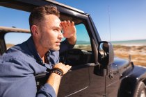 Homem caucasiano pensativo sentado no carro à beira-mar protegendo os olhos do sol e admirando a vista. viagem de verão e férias na natureza. — Fotografia de Stock