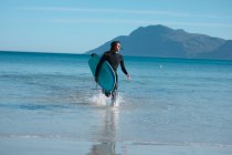 Homem com prancha de surf correndo enquanto espirra água na praia durante o dia ensolarado. hobbies e esporte aquático. — Fotografia de Stock