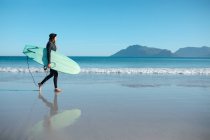 Vista lateral del surfista que lleva la tabla de surf caminando en la orilla mientras mira el cielo azul con espacio para copiar. pasatiempos y deportes acuáticos. - foto de stock