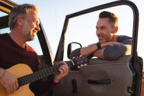 Feliz casal gay caucasiano tocando guitarra e rindo, saindo de carro à beira-mar. viagem de verão e férias na natureza. — Fotografia de Stock