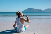 Rückansicht eines Mannes, der Wasser aus der Flasche trinkt, während er an sonnigen Tagen am Strand Yoga macht. Erfrischung und Fitness mit gesundem Lebensstil. — Stockfoto