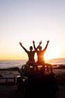 Rear view of caucasian gay male couple sitting on car roof raising arms at sunset by the sea. summer road trip and holiday in nature. — Stock Photo