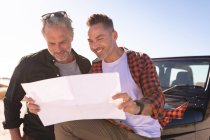 Happy caucasian gay male couple reading map, smiling by a car at seaside. summer road trip and holiday in nature. — Stock Photo