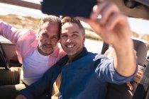 Happy caucasian gay male couple taking selfies sitting in car at seaside. summer road trip and holiday in nature. — Stock Photo