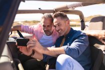 Happy caucasian gay male couple taking selfies sitting in car at seaside. summer road trip and holiday in nature. — Stock Photo