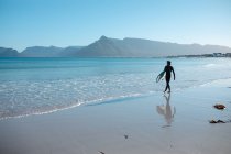 Hombre surfista caminando a lo largo de la orilla con tabla de surf contra el cielo azul claro y espacio de copia. pasatiempos y deportes acuáticos. - foto de stock