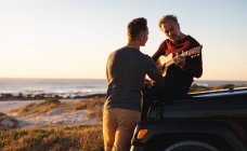 Heureux couple gay caucasien mâle traînant sur la plage au coucher du soleil, assis sur la voiture à jouer de la guitare. road trip d'été et vacances dans la nature. — Photo de stock