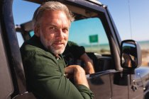 Retrato de un hombre caucásico feliz sentado en coche en la playa en un día soleado. viaje por carretera de verano y vacaciones en la naturaleza. - foto de stock