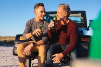 Happy caucasian gay male couple drinking bottles of beer, sitting on car at seaside. summer road trip and holiday in nature. — Stock Photo