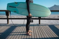 Bassa sezione di uomo che trasporta tavola da surf mentre in piedi sulla tavola da pavimento in spiaggia durante la giornata di sole. hobby e sport acquatici. — Foto stock
