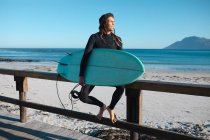 Чоловічий серфер, який несе дошку для серфінгу, дивлячись далеко, сидячи на дерев'яних перилах на пляжі. хобі і водні види спорту . — стокове фото