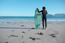 Vista posteriore dell'uomo in muta in piedi con tavola da surf guardando il cielo limpido con spazio copia dalla spiaggia. hobby e sport acquatici. — Foto stock