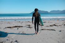 Вид сзади на человека в гидрокостюме, идущего с доской для серфинга на пляже к копировальному пространству на голубом небе. хобби и водные виды спорта. — стоковое фото