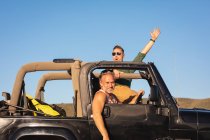 Portrait of happy caucasian gay male couple raising arms sitting in car on sunny day at seaside. summer road trip and holiday in nature. — Stock Photo