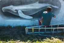 Vista trasera del artista independiente masculino sentado en la escalera haciendo hermosa pintura mural de ballena en la pared. arte urbano y habilidad. - foto de stock