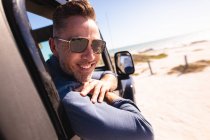 Портрет улыбающегося кавказца в солнцезащитных очках, сидящего в машине на берегу моря. летняя поездка и отдых на природе. — стоковое фото
