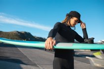 Hipster männliche Surfer mit Hut und Neoprenanzug tragen Surfbrett an sonnigen Tagen auf der Straße. Hobbys und Wassersport. — Stockfoto