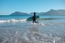 Uomo che trasporta tavola da surf in esecuzione mentre spruzza acqua sulla riva contro il cielo blu con spazio copia. hobby e sport acquatici. — Foto stock