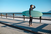 Male surfer carrying surfboard shielding eyes while standing on floorboard at beach. hobbies and water sport. — Stock Photo
