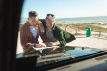 Glückliches kaukasisches schwules Paar beim Lesen einer Landkarte, angelehnt an die Motorhaube eines Autos am Meer. Sommer Roadtrip und Urlaub in der Natur. — Stockfoto