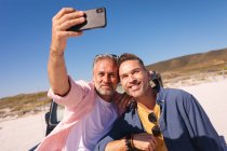 Feliz pareja gay caucásica abrazando y tomando selfies en un auto en la playa. viaje por carretera de verano y vacaciones en la naturaleza. - foto de stock
