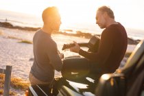 Heureux couple gay caucasien mâle traînant sur la plage au coucher du soleil, assis sur la voiture à jouer de la guitare. road trip d'été et vacances dans la nature. — Photo de stock