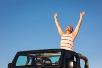 Happy caucasian gay male couple raising arms sitting in car on sunny day at seaside. summer road trip and holiday in nature. — Stock Photo