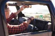 Homme caucasien réglage rétroviseur dans la voiture au bord de la mer. road trip d'été et vacances dans la nature. — Photo de stock