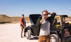 Stressed caucasian gay male couple talking on smartphone by broken down car at seaside. summer road trip and holiday in nature. — Stock Photo