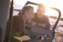 Happy caucasian gay male couple playing guitar and laughing, hanging out by car at seaside. summer road trip and holiday in nature. — Stock Photo