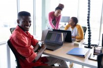 Портрет усміхненого афроамериканського бізнесмена з ноутбуком за столом у творчому офісі. креативний бізнес, бездротові технології та офісне робоче місце . — стокове фото